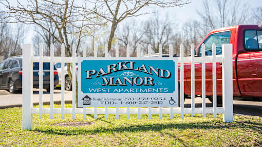 A sign for Parkland Manor West Apartments is displayed in front of a red truck.
