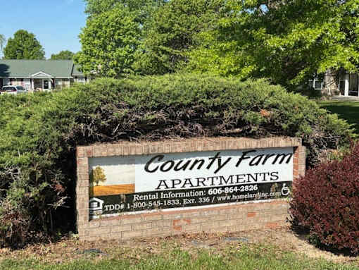 A sign for County Farm Apartments is surrounded by greenery.