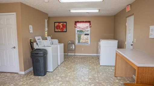 A room with a white fridge, a white washing machine, a white dishwasher, a trash bin, a wooden desk and a painting on the wall.