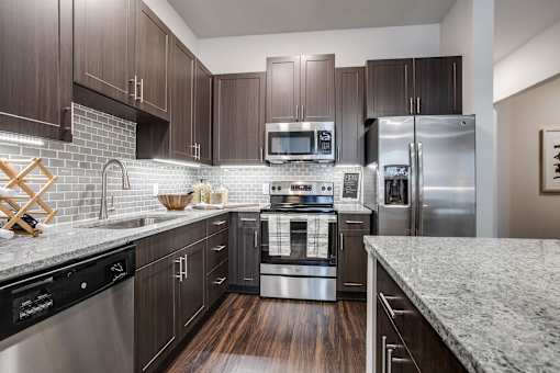 Modern kitchen with stainless steel appliances, tile backsplash, large island, granite countertops, and dark wood cabinetry.