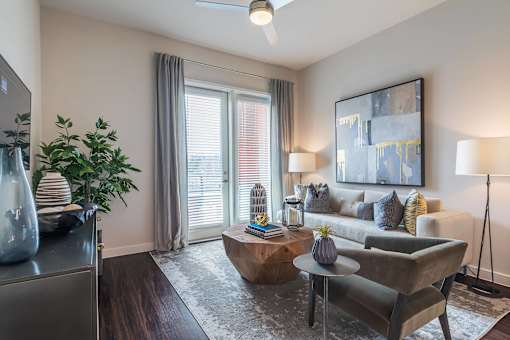 Modern apartment living room with comfortable seating, large windows, wood-style flooring, and abundant natural light.