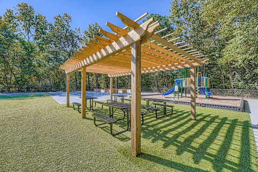 A wooden pergola with benches is situated in a grassy area with a playground in the background.