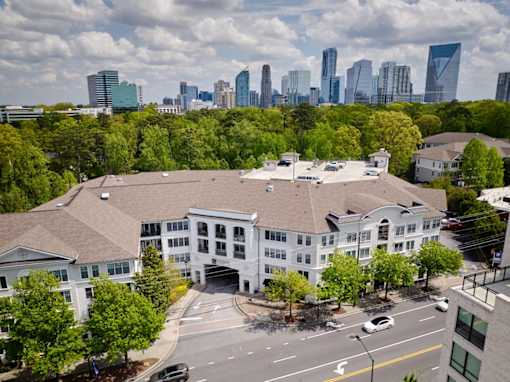 A large white building with a brown roof is surrounded by trees and has a city skyline in the background.