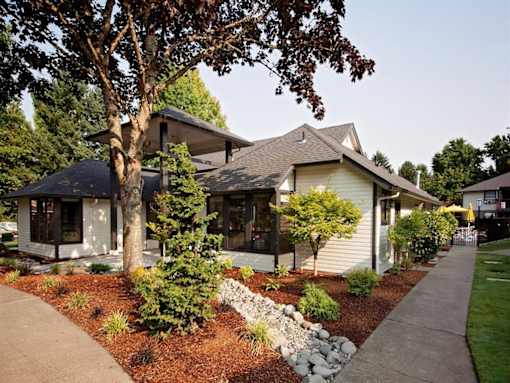 A house with a white siding and a brown roof is surrounded by a garden with a variety of plants and trees.