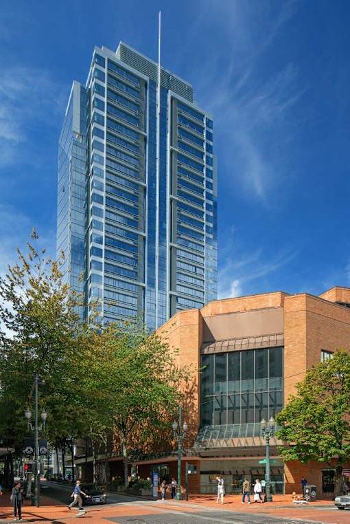 A modern glass building stands tall next to a smaller brick building with people walking around on the sidewalk.