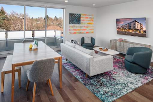 A living room with a grey couch, a wooden dining table and a painting of a building on the wall.