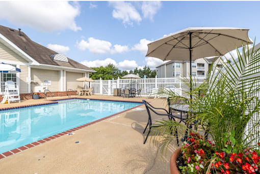 A pool surrounded by a white fence and a patio with chairs and an umbrella.