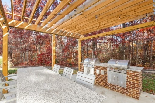 A wooden pergola with a grill and chairs is set up in a backyard.