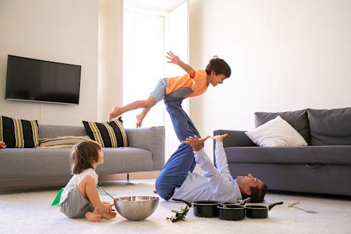 A man is doing a handstand while a child is sitting on the floor and another child is standing on his back.