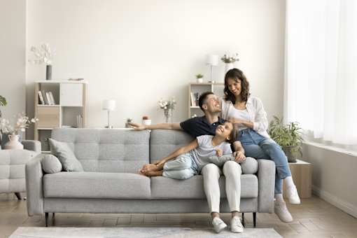 A woman and a child are sitting on a grey couch with a man lying on the floor.