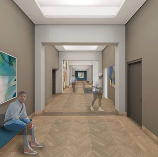 A man is sitting on a bench in a hallway with a person walking in the distance.