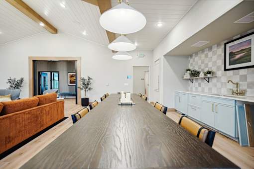 A long wooden table is in the middle of a room with a white ceiling and a painting hanging on the wall.