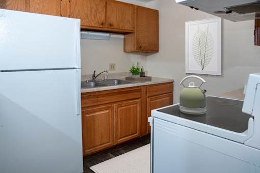 A white refrigerator stands next to a kitchen counter with a sink and a green kettle on top.