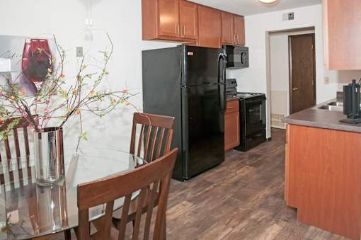 A black refrigerator sits in a kitchen with wooden cabinets and a glass table.