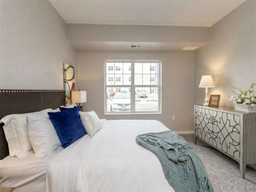 A bedroom with a large bed, a nightstand, and a window with a view of the street at The Barnum Apartments, White Bear Lake, Minnesota