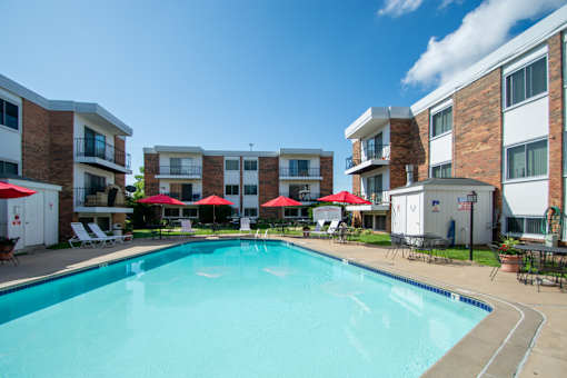 A swimming pool surrounded by red umbrellas and lounge chairs in front of apartment buildings.