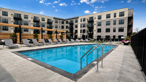 A swimming pool is surrounded by lounge chairs in front of apartment buildings.