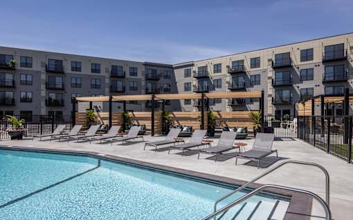 A swimming pool is surrounded by lounge chairs in front of apartment buildings.