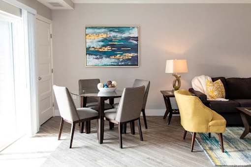 A living room with a grey couch, a yellow chair, a grey dining table and chairs, a painting on the wall, and a lamp on the side table.