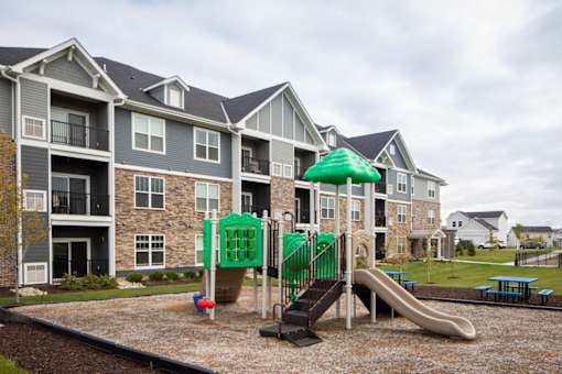 A playground with a green umbrella and slide in front of apartment buildings.