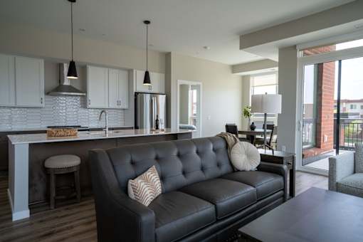 A black leather couch with a white pillow is in the middle of a kitchen and living room.