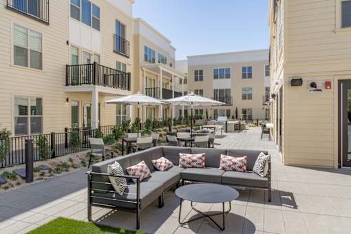 A patio with a couch, chairs, and umbrellas is surrounded by apartment buildings.