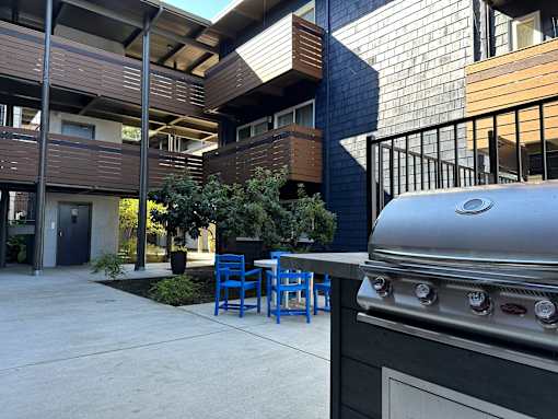 A silver grill is in the foreground of a modern building with blue chairs and tables.
