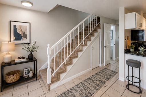 A well-lit foyer with a staircase, a bar stool, and a framed artwork on the wall.