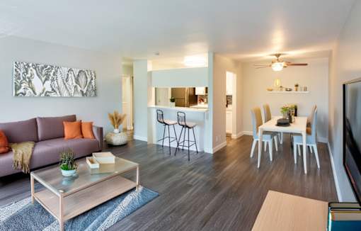 A modern living room with a grey sofa, a glass coffee table, and a dining area in the background.
