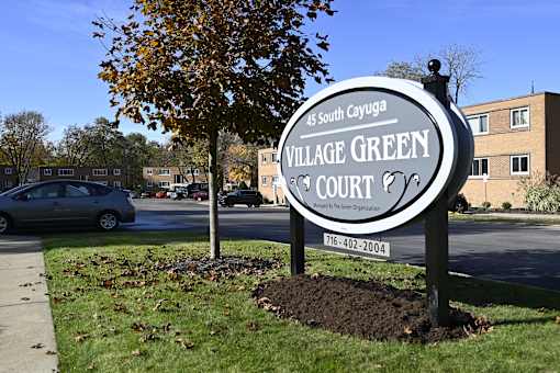 A sign for Village Green Court stands in front of a tree and a building.