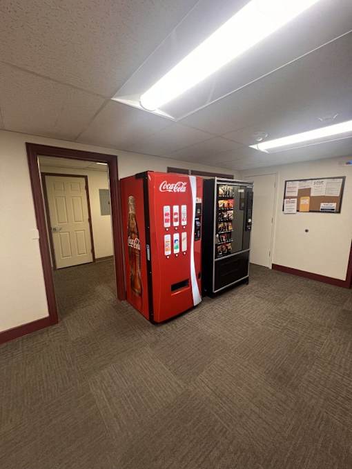 A Coca-Cola vending machine sits in a room with carpeted flooring and white walls.
