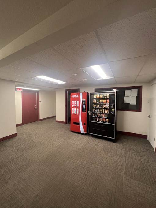A Coca-Cola vending machine is in the middle of a carpeted room.