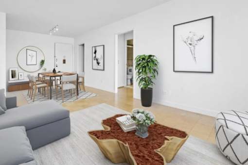 A living room with a grey sofa, a coffee table with a vase of flowers, a potted plant, and pictures on the wall at The Caldwell Apartments, New York, NY, 10028