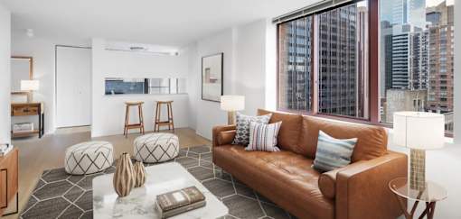 A living room with a brown leather couch, a white coffee table, and a large window overlooking the city at The Ellington Apartments, New York, NY