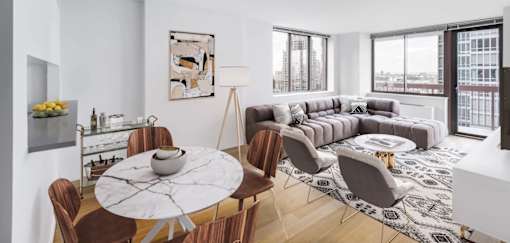 A modern living room with a large grey sofa, a round marble table, and a view of the city at The Ellington Apartments, New York, 10019