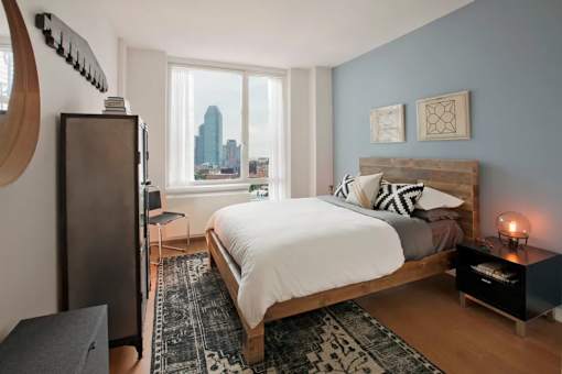 A bedroom with a large bed, a TV, and a city view through the window at The Maximilian Apartments, Long Island City
