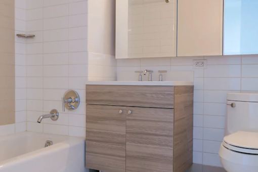 A white toilet sits next to a sink with a wooden cabinet above it at The Maximilian Apartments, Long Island City, NY, 11101