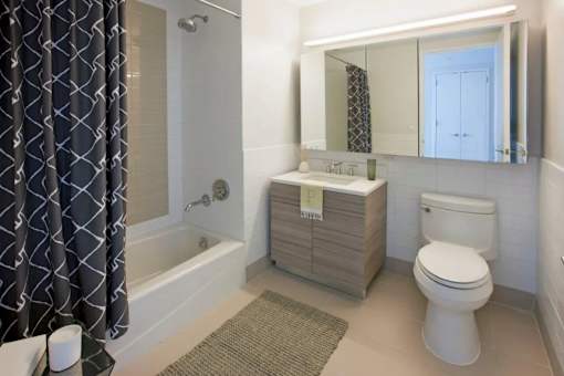 A bathroom with a white toilet, a sink, and a bathtub with a grey shower curtain at The Maximilian Apartments, Long Island City, New York