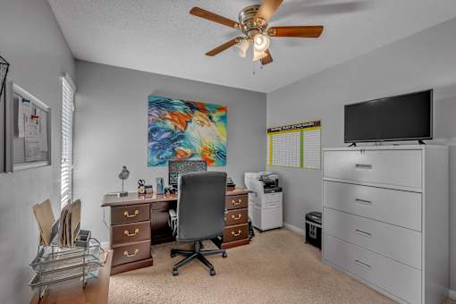 A room with a ceiling fan, a desk with a chair, a filing cabinet, and a television.