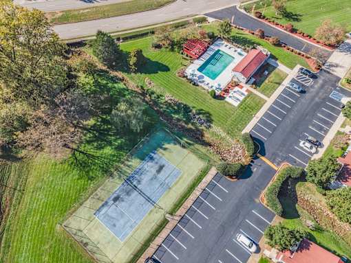 A tennis court is surrounded by a parking lot and a grassy area.