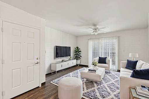 A living room with a white couch, a white chair, a white ottoman, a white rug, a white ceiling fan, a white door, a white cabinet, a white television, and a white wall.