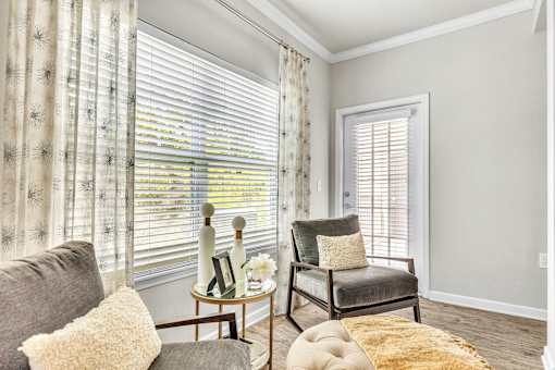 A living room with a grey couch, a chair, a small table with a vase and a picture frame on it, and a window with white blinds.