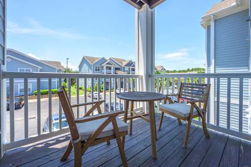 A wooden table and two chairs are on a deck with a view of a street and houses.
