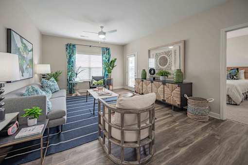 A well-lit, modern living room with a grey sofa, a round coffee table, and a bed in the background at Waterleaf at Neely Ferry Apartments, South Carolina
