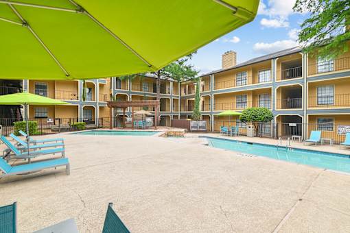 A large outdoor pool area with sun loungers and umbrellas in front of apartment buildings.