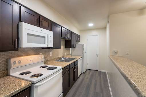 A kitchen with dark wood cabinets and white appliances in a 1 bedroom, 1 bath C, 653 sq ft apartment home with hardwoodlike flooring throughout at The Biltmore Apartments located in the Vickery Midtown neighborhood of Dallas, TX.