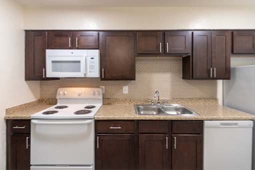 A kitchen with dark wood cabinets and white appliances in a 1 bedroom, 1 bath C, 653 sq ft apartment home with hardwoodlike flooring throughout at The Biltmore Apartments located in the Vickery Midtown neighborhood of Dallas, TX.