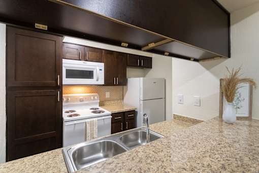 A kitchen with dark wood cabinets and white appliances in a 1 bedroom, 1 bath B, 629 sq ft apartment home at The Biltmore Apartments located in the Vickery Midtown neighborhood of Dallas, TX.