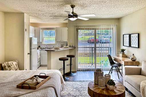 Living and sleeping area in an Efficiency apartment at Cambridge Court Apartments in Dallas, Texas, showing carpeted flooring, patio door, dining space, and connection to the kitchen.