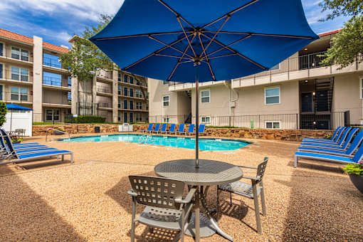 A pool area with lounge chairs, patio tables and chairs with blue umbrellas and stainless steel BBQ in front of apartment buildings at Princeton Court Apartments in the Vickery Midtown neighborhood of Dallas, TX.